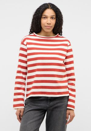 Femeie cu păr creț, purtând o bluză cu mâneci lungi, în dungi roșii și albe, și blugi gri închis, stând în picioare pe un fundal simplu.