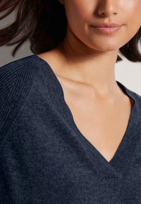 Frau trägt einen dunkelblauen V-Ausschnitt-Pullover, der das Schlüsselbein und einen Teil des Kinns und der Lippen zeigt, mit braunem Haar, das nach hinten fließt.