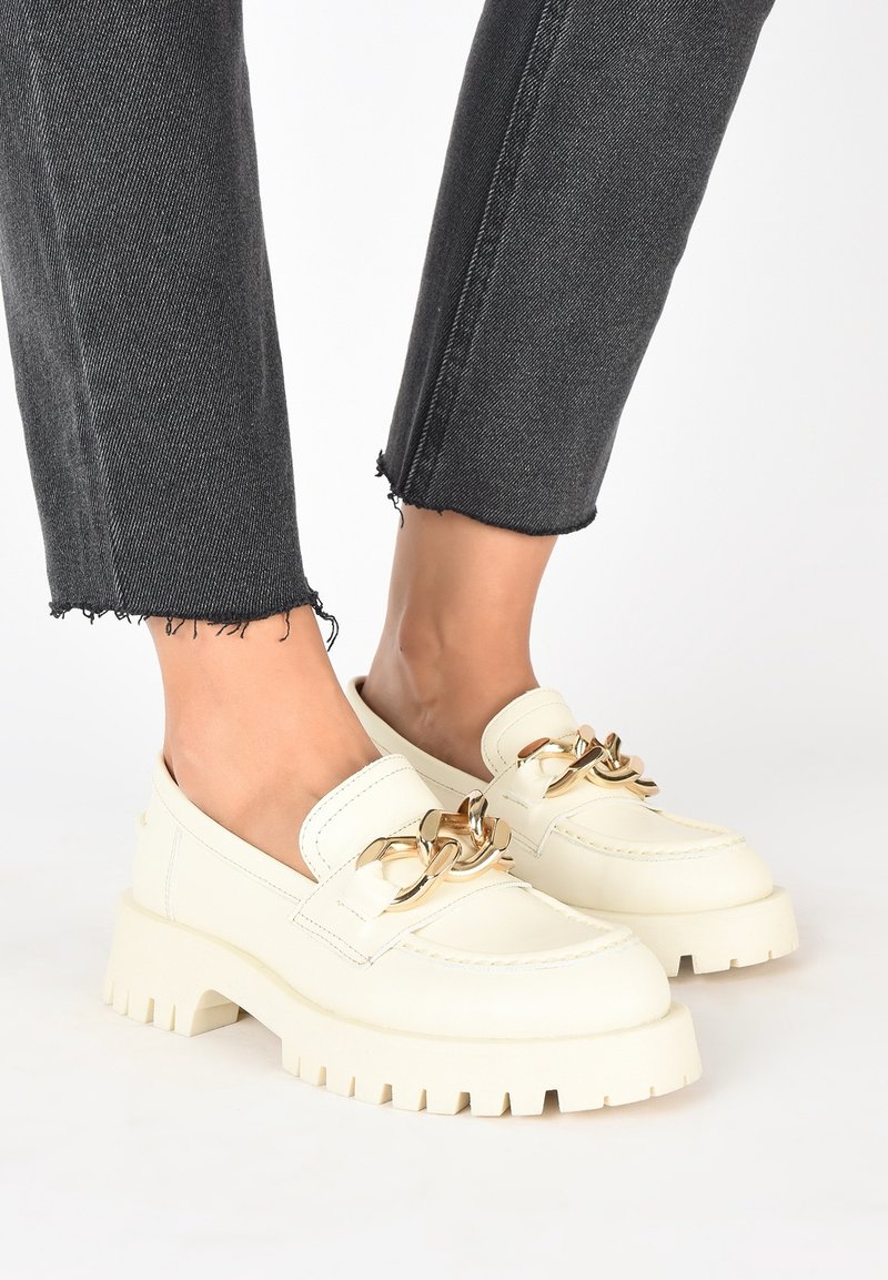 Chaussures mocassins épaisses crème avec un détail chaîne dorée surdimensionnée, portées avec un jean noir raccourci à ourlet effiloché pieds nus.