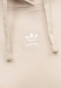 Μπεζ φούτερ με κουκούλα από απαλό ύφασμα, με λευκό κεντητό λογότυπο Adidas και κορδόνια. Ομαλή υφή με άνετη εφαρμογή.