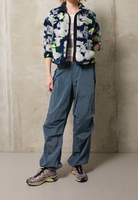 Kurtka polarowa z granatowym, białym i neonowym wzorem. Do tego lekkie, teksturowane niebieskie spodnie i szare buty sportowe.