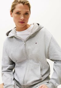 Felpa grigia con zip realizzata in tessuto morbido, dotata di una tasca frontale e un piccolo logo. L'interno presenta una fodera di un grigio chiaro contrastante.