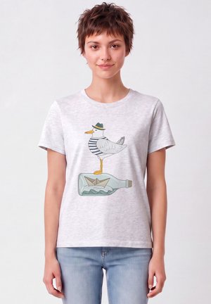 Jonge vrouw draagt een lichtgrijs T-shirt met een meeuw in een gestreept overhemd en hoed, staande op een fles met een papieren bootje erin.