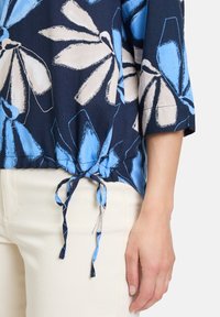 Marinebluse med store blå og hvide blomstermønstre, med en knyttet kant og trekvartærmer, parret med lyse beige bukser.