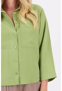 Groen, kortaf shirt gemaakt van gladde stof met een knoopsluiting en twee borstzakken. Eenvoudig ontwerp met een relaxte pasvorm.