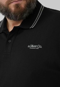 Schwarzes Polohemd aus strukturiertem Stoff, mit einem zweifarbigen Kragen mit grauen Streifen und einem silbernen Logo auf der Brust.