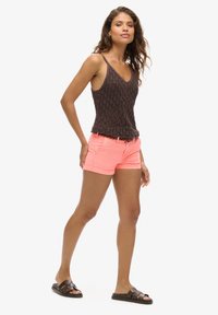 Top in maglia marrone scuro con orlo ondulato, abbinato a pantaloni corti di jeans rosa brillante. Modello che indossa sandali neri a scivolo, in posizione profilo.