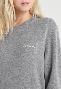 Grå stickad tröja med avslappnad passform, rund halsringning och uppvikta ärmar. Har en diskret vit logotyp "CALVIN KLEIN JEANS" på bröstet.