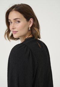 Black textured blouse with ruffled neckline and short, puffed sleeves. Features a subtle keyhole at the back and decorative trim.