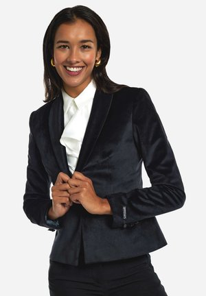Černý sametový blazer s přiléhavým střihem, saténovými klopami a dekoračními knoflíky. Spárovaný s bílou košilí s volánky pod ním.