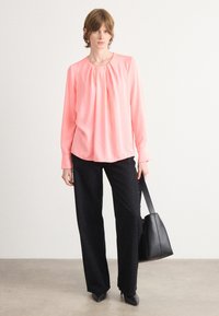 Korallfarbige Bluse mit langen Ärmeln und plissiertem Ausschnitt, kombiniert mit weiten schwarzen Hosen mit gesprenkelter Textur und einer schwarzen Handtasche.