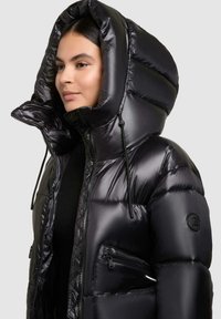 Vrouw in een glanzende zwarte pufferjack met een grote capuchon, ritszakken en een hoge kraag, tegen een effen lichte achtergrond.