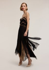 Abito nero a strati con bordatura in pizzo dorato, gonna trasparente fluente, spalline sottili e lunghezza fino alla caviglia. Abbinato a scarpe con tacco nude.