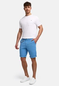 Man standing wearing a white short-sleeve t-shirt, light blue rolled-up shorts with a keychain, and white sneakers on a plain background.