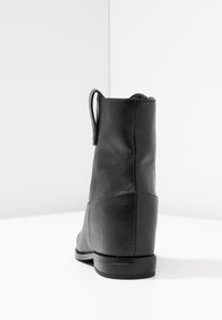 Bottine en cuir noir avec une boucle de tirage à l'arrière, semelle basse et coutures visibles, photographiée sur un fond blanc en coin.