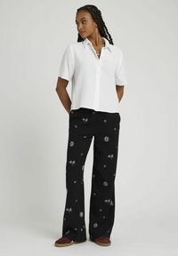 Chemise blanche à manches courtes, boutonnée, avec un tissu texturé, associée à un pantalon noir évasé orné de motifs de soleil et de palmiers blancs. Baskets rouges.