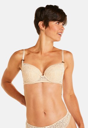 Soutien-gorge en dentelle beige avec des motifs floraux, soutien-gorge à armatures, bretelles réglables et un petit nœud au centre. Détail de tissu texturé.