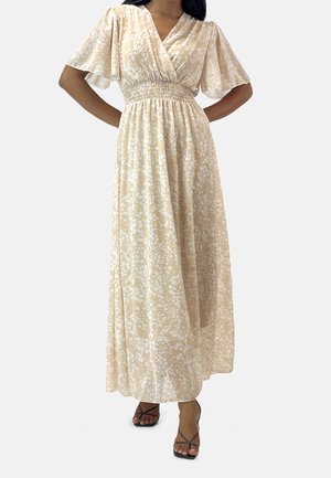 Femme portant une robe maxi beige à motifs floraux avec des manches volantées, une taille smockée et des sandales à talons hauts noires à lanières.