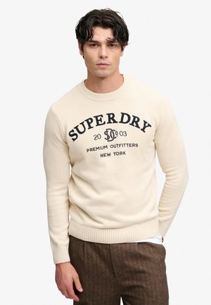 Mand med mørkt hår iført en cremefarvet sweater med teksten "Superdry Premium Outfitters New York" og brune bukser, stående med den ene hånd i lommen.