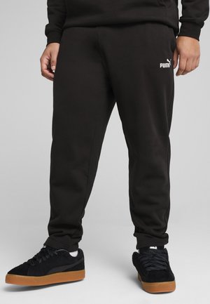 Sorte sweatpants lavet af en blanding af bomuldsstof, med et hvidt logo på venstre lår, parret med sorte sneakers i ruskind og gummisål.