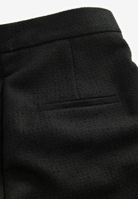 Tissu noir texturé avec une poche passepoilée horizontale à l'arrière et une ceinture, montrant une partie de la section arrière d'un vêtement.