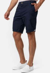 Shorts en coton bleu marine avec ourlets retroussés, avec poches latérales et fermeture à bouton marron. Portés avec des chaussures blanches à enfiler.