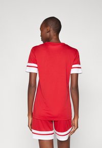 Rode sportieve top en shorts van gladde stof, met witte strepen op de mouwen en shorts. De achterkant laat een relaxte pasvorm zien.