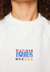 Weißes Sweatshirt mit geripptem Rundhalsausschnitt, bestickt mit "HILFIGER 1985 N.Y.C. U.S.A" in Rot, Blau und Gelb.