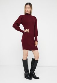 Burgundy ribbed knit dress with high neck, puff sleeves, and a fitted silhouette, paired with knee-high black leather block-heeled boots.
