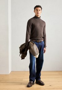 Rjava turtleneck pulover, temno modri traper hlače, rjav pas, drži olivno zeleno jakno s kremasto podlogo in rjave loafers čevlje.