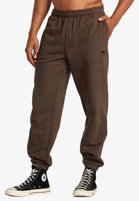 Braune Sweatpants mit einem elastischen Bund und Seitentaschen, versehen mit einem kleinen schwarzen Markenlogo auf der Hüfte. Kombiniert mit schwarzen High-Top-Sneakern.