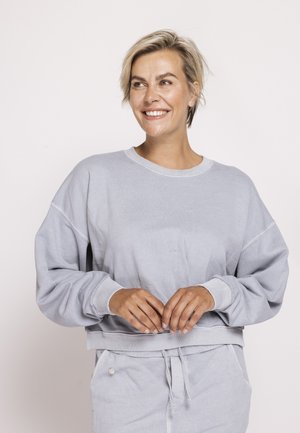 Woman with short blonde hair wearing a light gray oversized sweatshirt and matching pants, smiling and looking to the side.