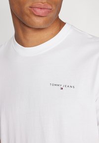 Fehér pamut póló kerek nyakú kivitelben, kis fekete "TOMMY JEANS" logóval, amely alatt színes akcentus található.