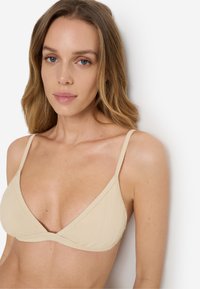 Νεαρή γυναίκα με ανοιχτά καστανά μαλλιά, που φοράει μπεζ τριγωνικό μπικίνι με υφή, πάνω σε απλό λευκό φόντο.