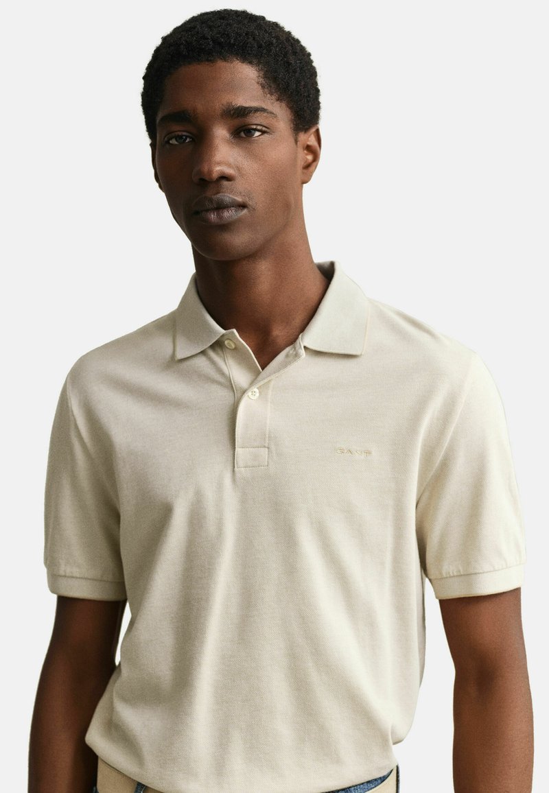 Jonge man die een beige polo met korte mouwen draagt met een klein logo op de borst, staand tegen een effen lichte achtergrond.