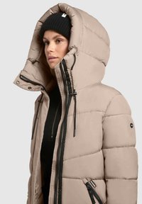 Bézs színű steppelt dzseki nagy kapucnival, fekete díszítésekkel, cipzáras zsebekkel és steppelt mintázattal. Az anyag sima és szigetelt hatású.