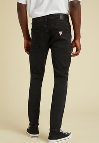 Uomo vestito con jeans neri slim-fit con tasche posteriori e una maglietta bianca, in piedi su uno sfondo beige.