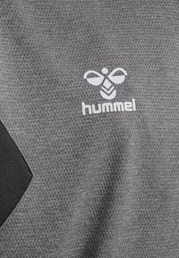 Szara, teksturowana tkanina z czarnym trójkątnym wzorem oraz białym logo Hummel przypominającym pszczołę, znajdującym się w górnym centrum.