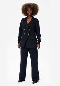 Zwarte dubbele blazer met gouden knopen, slim fit, en twee voorzakken, gecombineerd met wijde zwarte broek en puntige zwarte schoenen.