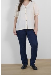 Blouse créme avec des manches bouffantes et un décolleté en V, ornée de boutons sur le devant. Associée à un jean slim bleu marine et à des mocassins noirs sur un sol en bois.