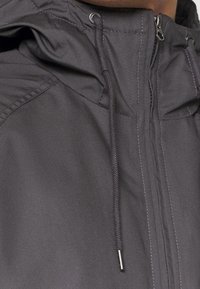 Acercamiento a una chaqueta con capucha de color gris oscuro, con cremallera y cordones, parcialmente cerrada cerca del cuello.