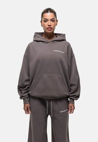 Grijze oversized hoodie, zachte stof, kangoeroezak, witte logo op de borst. Gepaard met bijpassende broek, voorzien van een tailleband met trekkoord en logo.