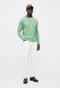 Zeleni pleteni džemper, bijele traperice i smeđa kapa. Džemper ima okrugli izrez i rebraste manžete. Crne cipele upotpunjuju odjevnu kombinaciju.