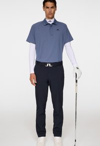 Blauwe polo met korte mouwen, witte onderlaag, zwarte riem met logo, marineblauwe broek en witte golfhandschoenen, terwijl een golfclub wordt vastgehouden.