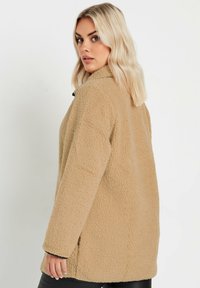 Beige fleece-takki, jossa on teksturoidun pinnan, pitkät hihat ja kaulus. Ominaisuuksiin kuuluu rento istuvuus ja sivutaskut mukavuutta varten.