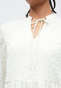 Γυναίκα με λευκό πουκάμισο με ανοιχτό γκρι πουά και δέσιμο σε φιόγκο στο λαιμό, εμφανίζοντας πλεξούδα στα μαλλιά και άνω κορμό.