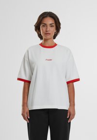 T-shirt bianco oversize con colletto e polsini a coste rossi; presenta testo ricamato sul petto; realizzato in tessuto morbido. Pantaloni neri visibili.