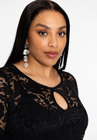 Zwart kanten jurk met een sleutelgat-decolleté. Lang haar en kristal hangoorbellen voegen detail toe. Zachte huid en natuurlijke make-up accentueren de look.