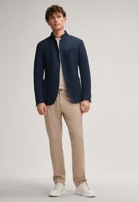 Marineblazer mit Stehkragen, vorderem Knopfverschluss und zwei Taschen. Kombiniert mit hellbeigen Hosen und weißen Sneakern. Glatte Textur.