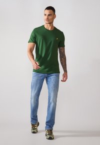 Grønn t-skjorte med kort erme og rund hals, med en liten gul logo. Båret med lyseblå jeans og joggesko med camo-mønster.